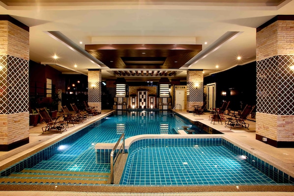 A large indoor swimming pool with a tiled floor and walls, surrounded by lounge chairs and a reception area with a check-in counter.