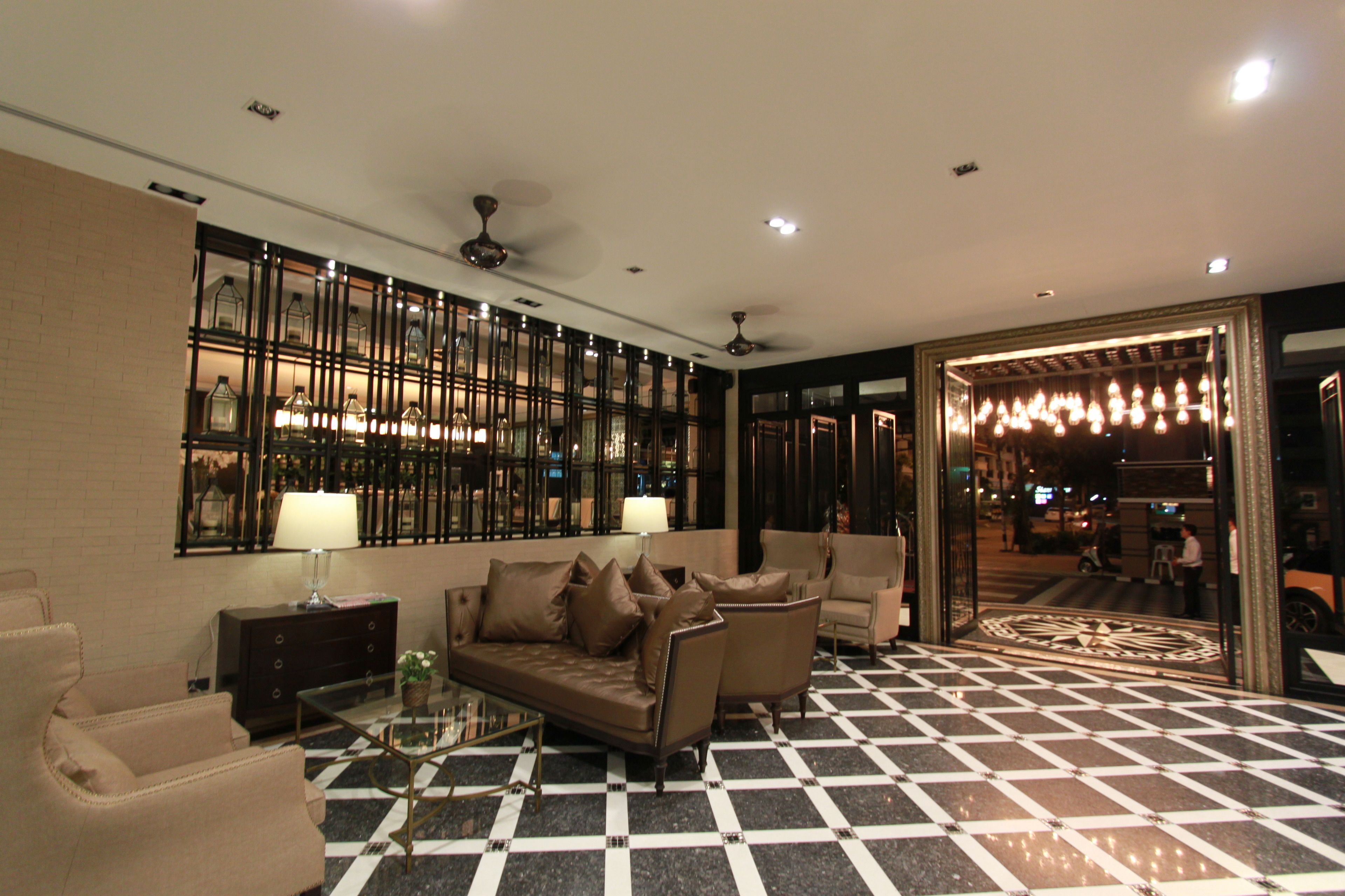 A lobby with a checkered floor, a sofa, armchairs, a glass coffee table, a lamp, and a wall with framed pictures.