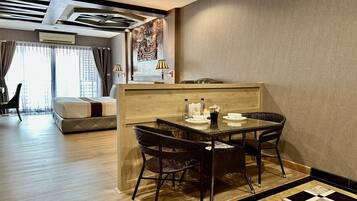 Habitación de hotel con comedor que incluye un separador de madera, dos sillas de mimbre, una mesa de vidrio y una cama con cabecera.
