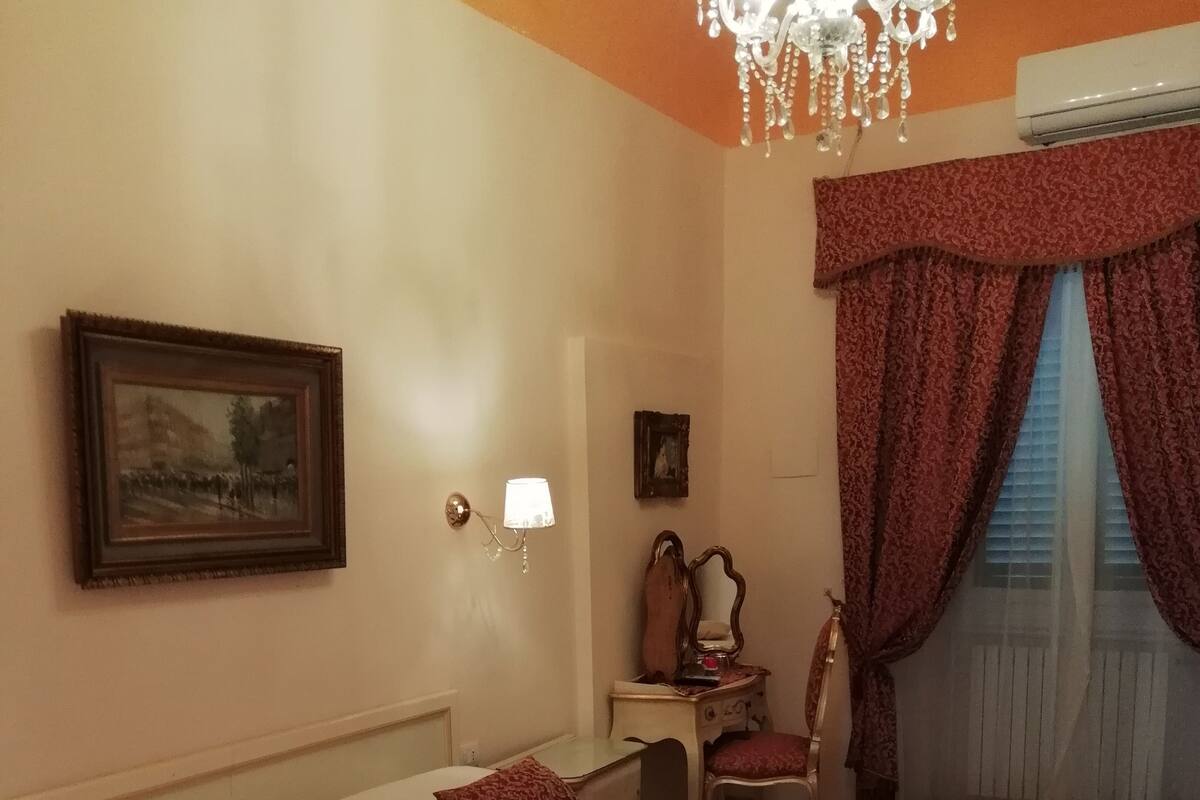 A hotel room with a large bed, a desk, a chair, a chandelier, and a painting on the wall.