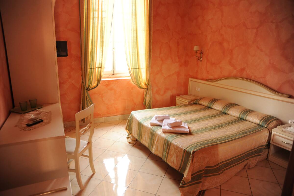 A hotel room with a bed, a desk, a chair, a nightstand, and a window with curtains.