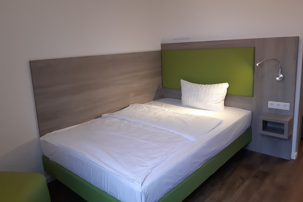 Uma cama de solteiro com roupa de cama branca, cabeceira verde e mesa de cabeceira com abajur.