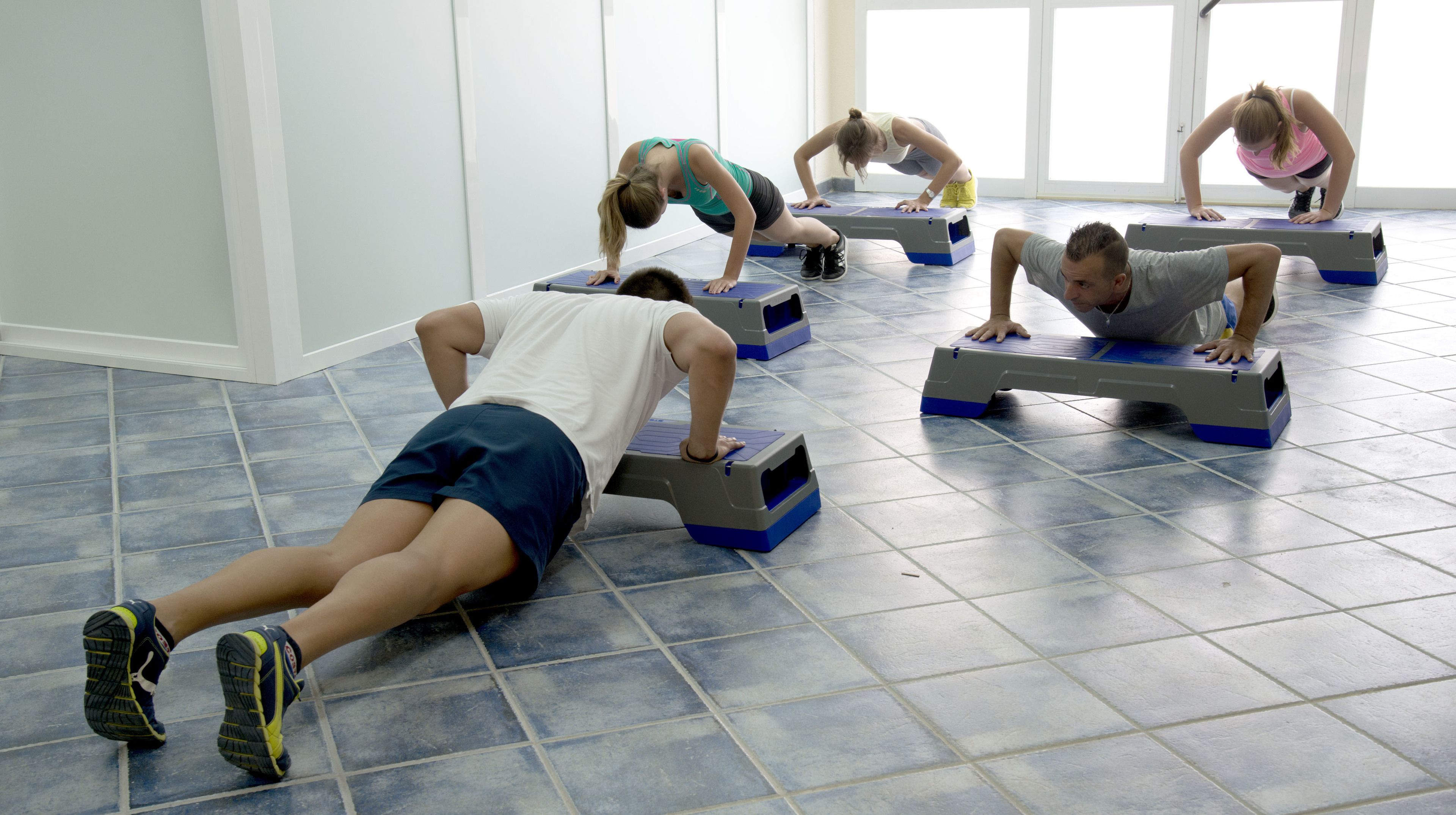Un grupo de personas haciendo flexiones sobre plataformas de ejercicio azul y gris en una habitación con suelo de baldosas y ventanas amplias.