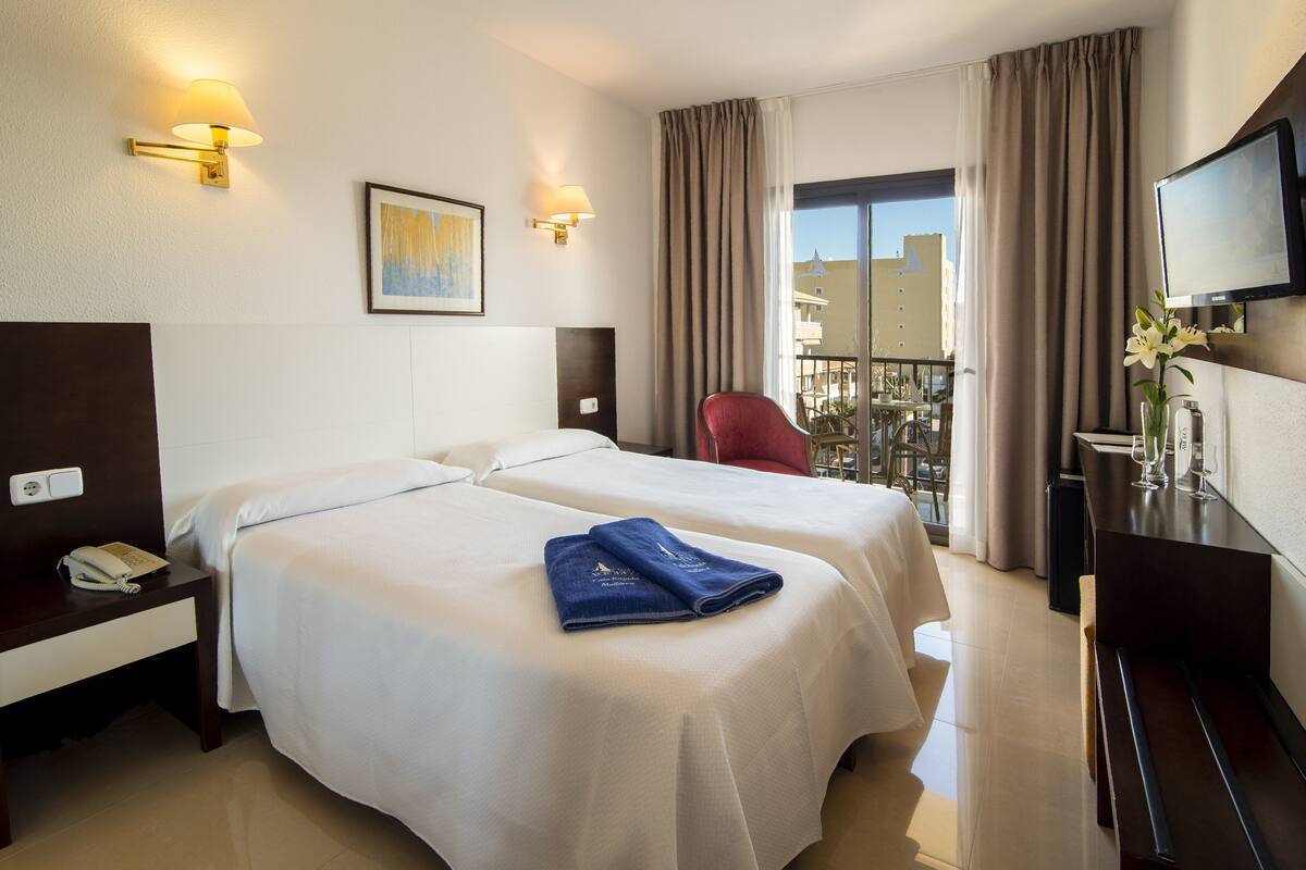 A hotel room with two beds, a desk with a telephone, a TV, and a balcony with a view of buildings.