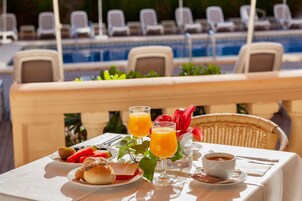 Una mesa dispuesta con alimentos para el desayuno, como cruasanes, fruta y jugo de naranja, con vista a una piscina y sillones de descanso.