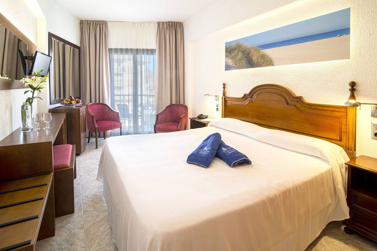 A hotel room with a large bed, a desk with a computer, two chairs, and a window with curtains.