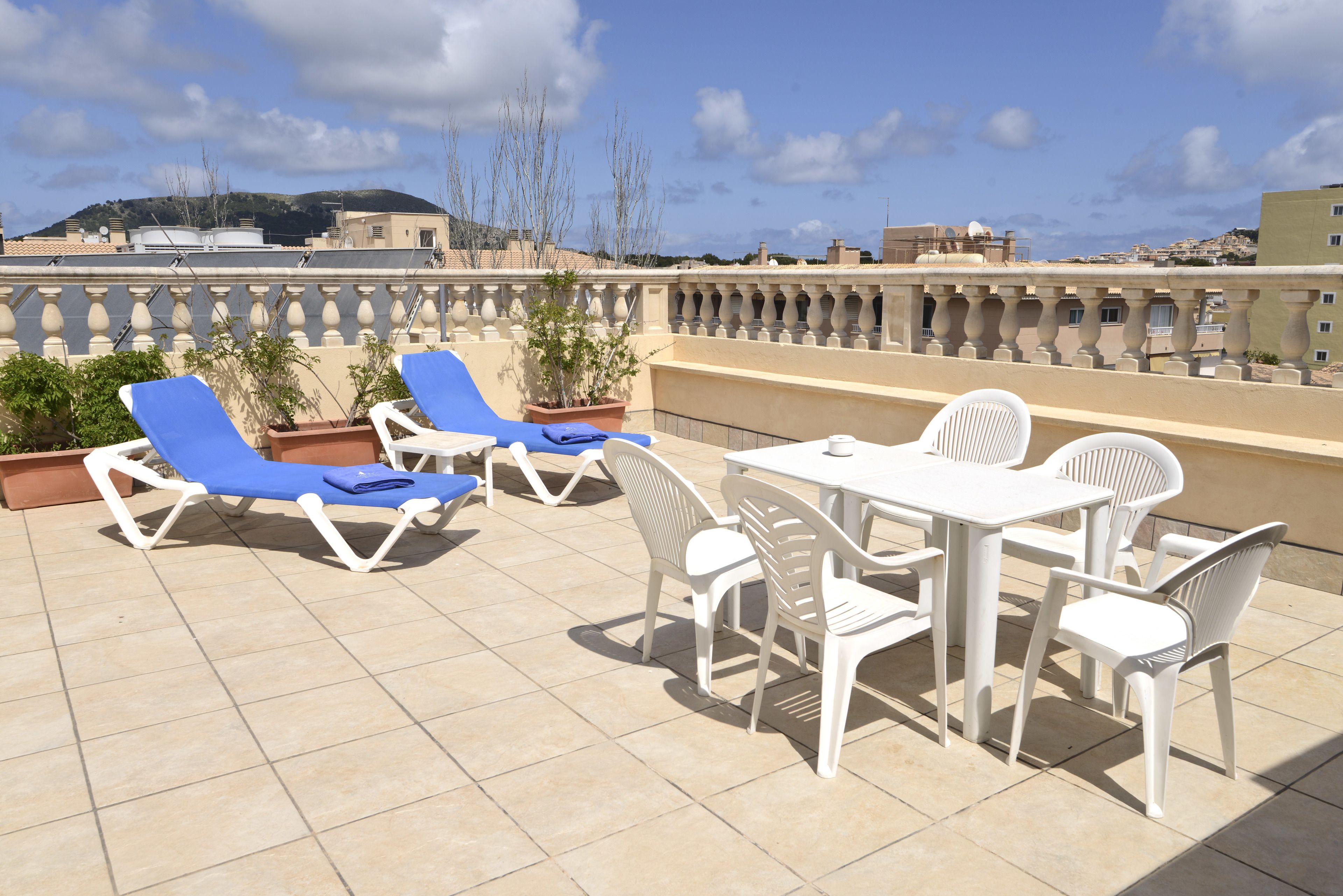 A rooftop terrace with blue and white lounge chairs, a table, and chairs, surrounded by potted plants.