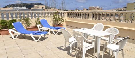 A rooftop terrace with blue and white lounge chairs, a table, and chairs, surrounded by potted plants.