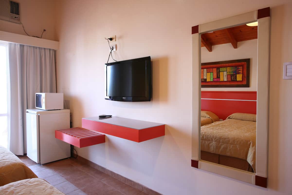 A hotel room with a TV mounted on the wall, a small refrigerator, a red shelf, and a mirror reflecting a bed.