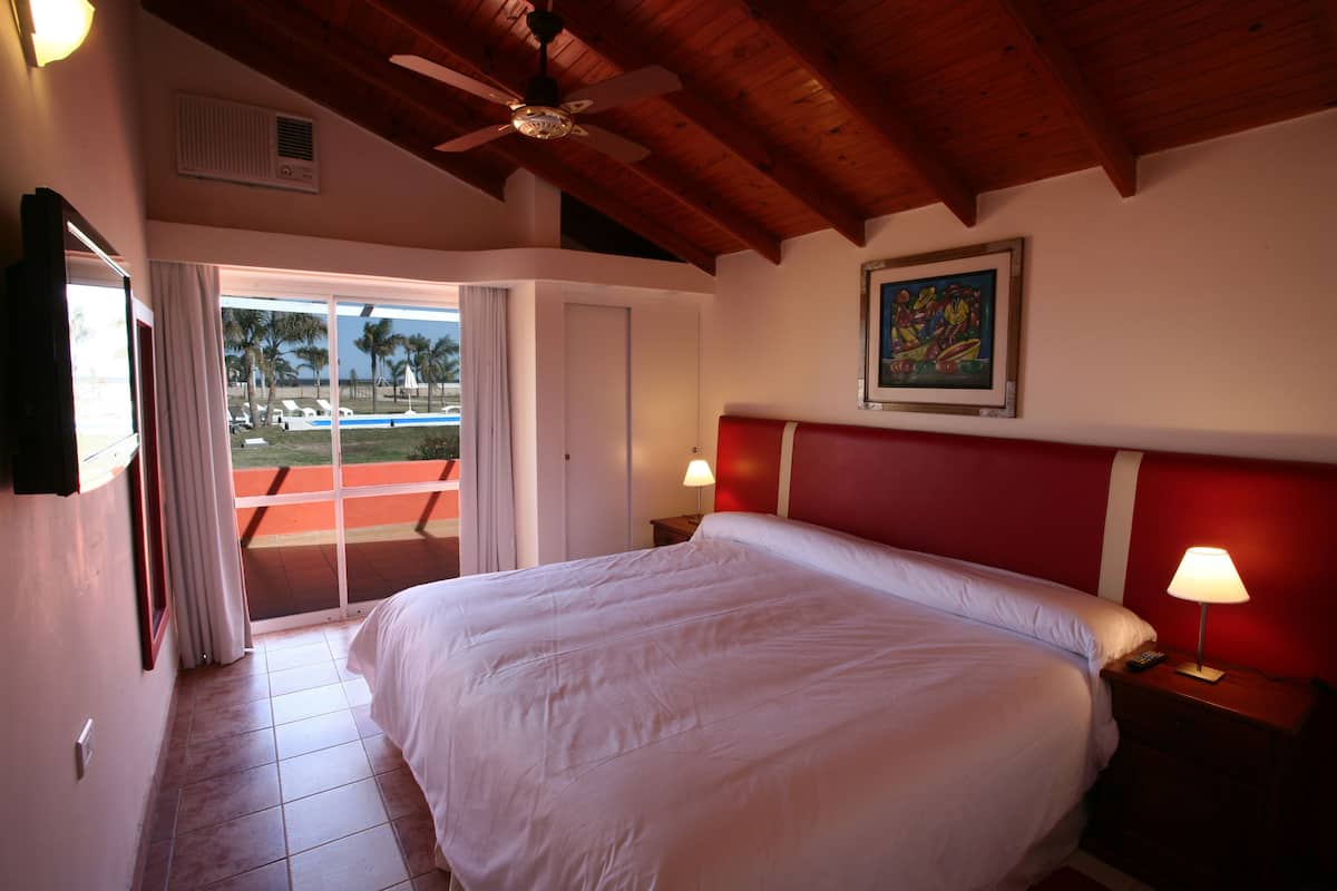 A hotel room with a large bed, a ceiling fan, a painting on the wall, and a view of the pool through the sliding door.
