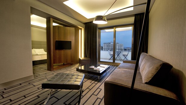 Ein Hotelzimmer mit Bett, Sofa, Couchtisch, Fernseher und Blick auf die Stadt durch große Fenster.