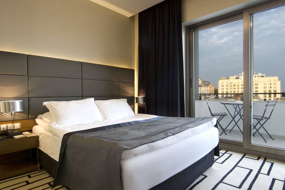 A modern hotel room with a large bed, a nightstand, a desk, and a view of the city through large windows.