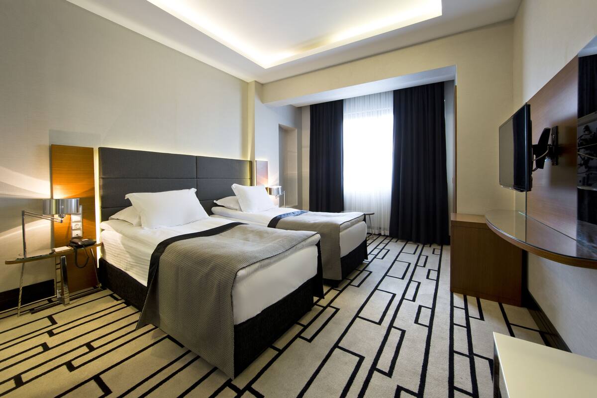 A hotel room with two beds, a TV, a desk, and a large window with curtains.