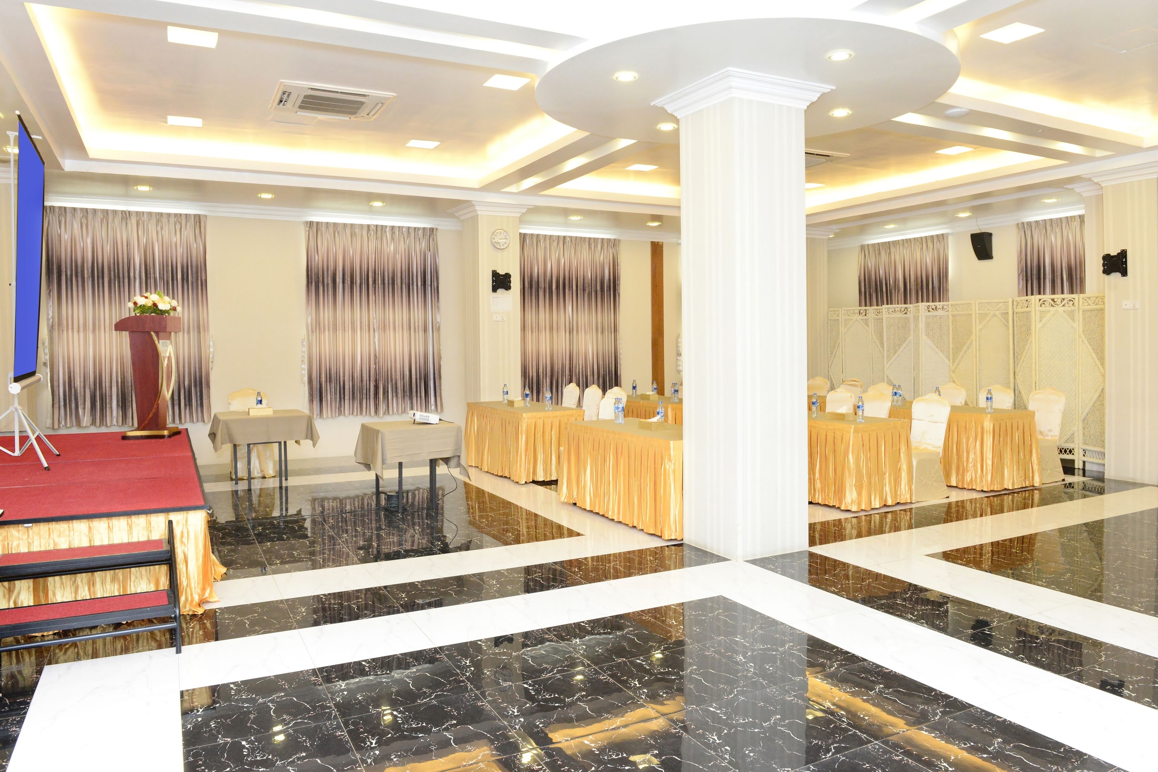 Un salón de conferencias con escenario, mesas y sillas, que destaca por su suelo de mármol pulido y un diseño de techo moderno.