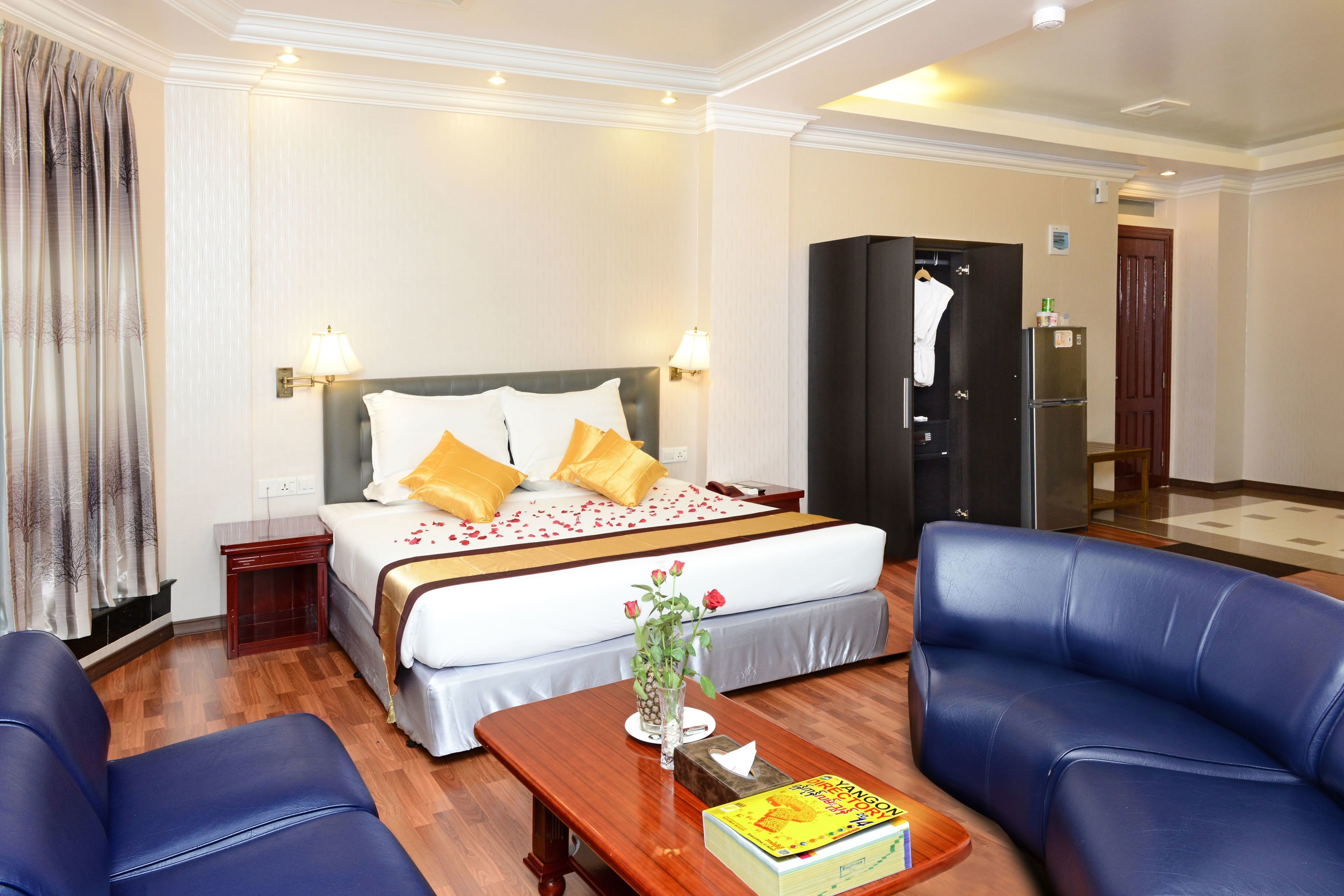 Una habitación de hotel con una cama, dos mesitas de noche, un pequeño escritorio, una mesa de centro de madera, un sofá azul y un armario negro.