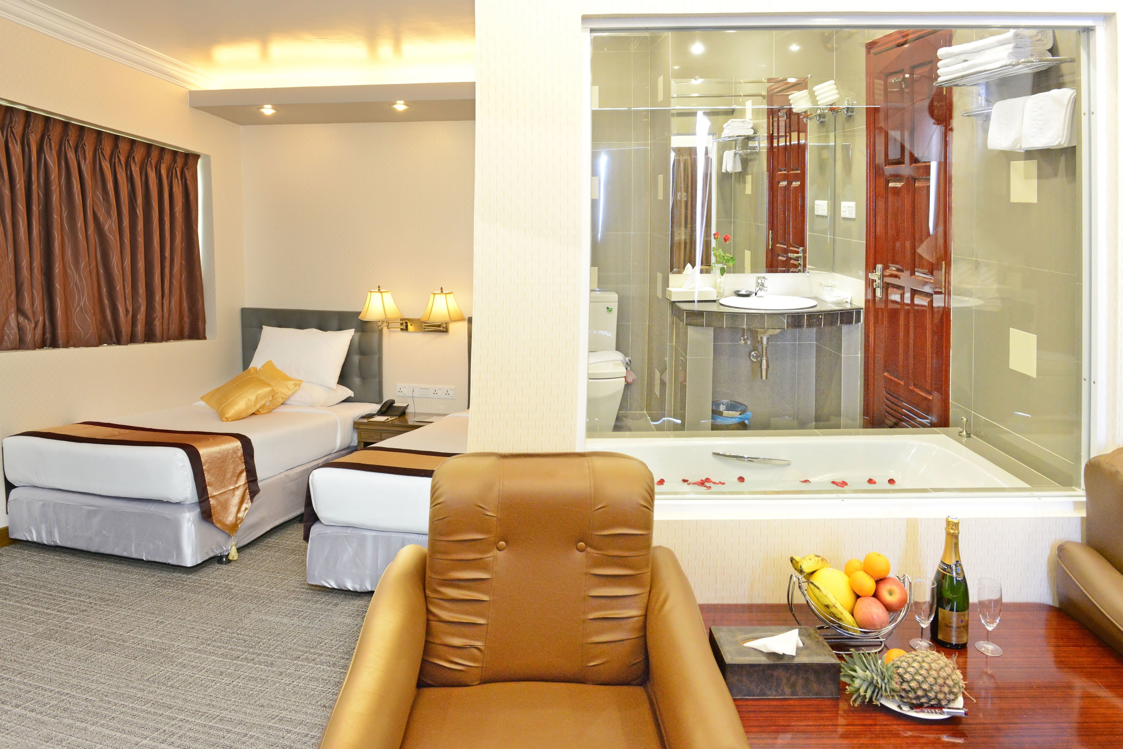 A hotel room with a bed, a chair, a table with fruit and a bottle, and a bathroom visible through a glass wall.
