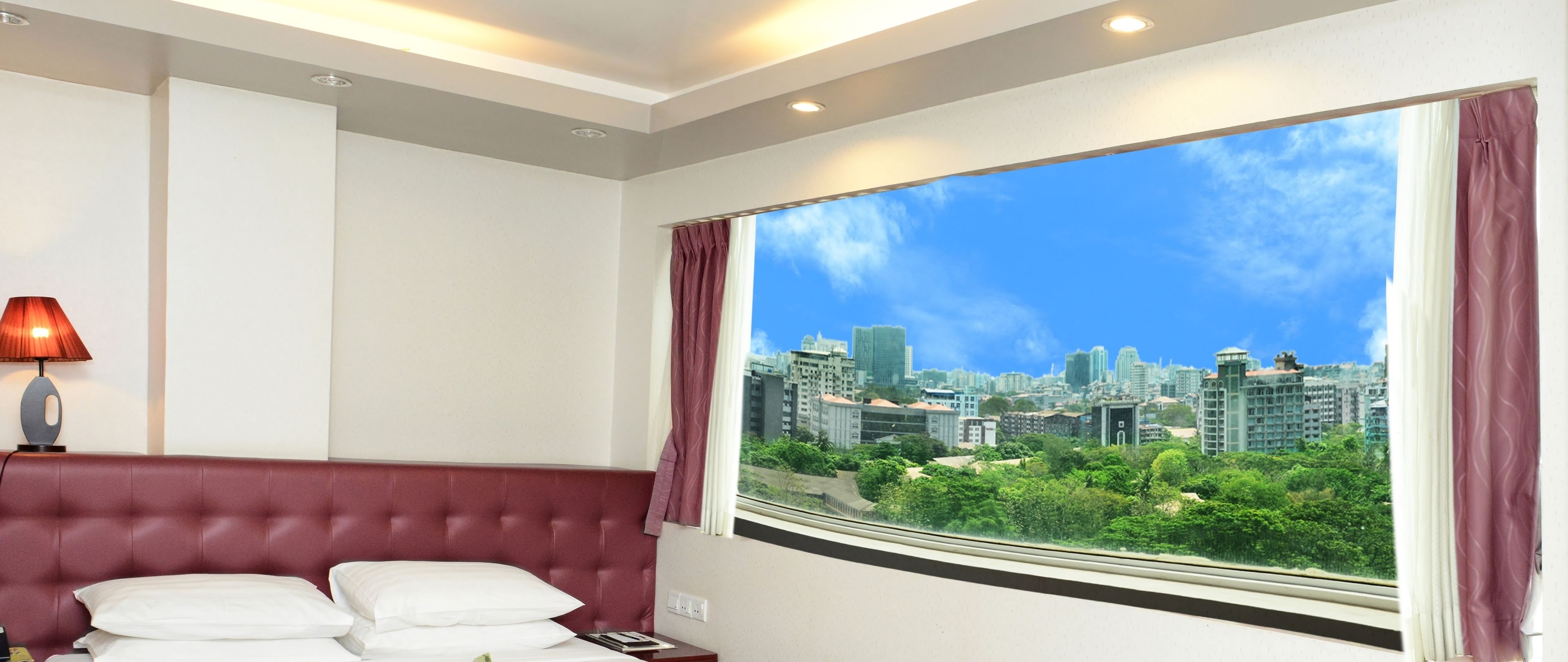 A hotel room with a large window offering a cityscape view, a red upholstered headboard, and a bedside lamp.