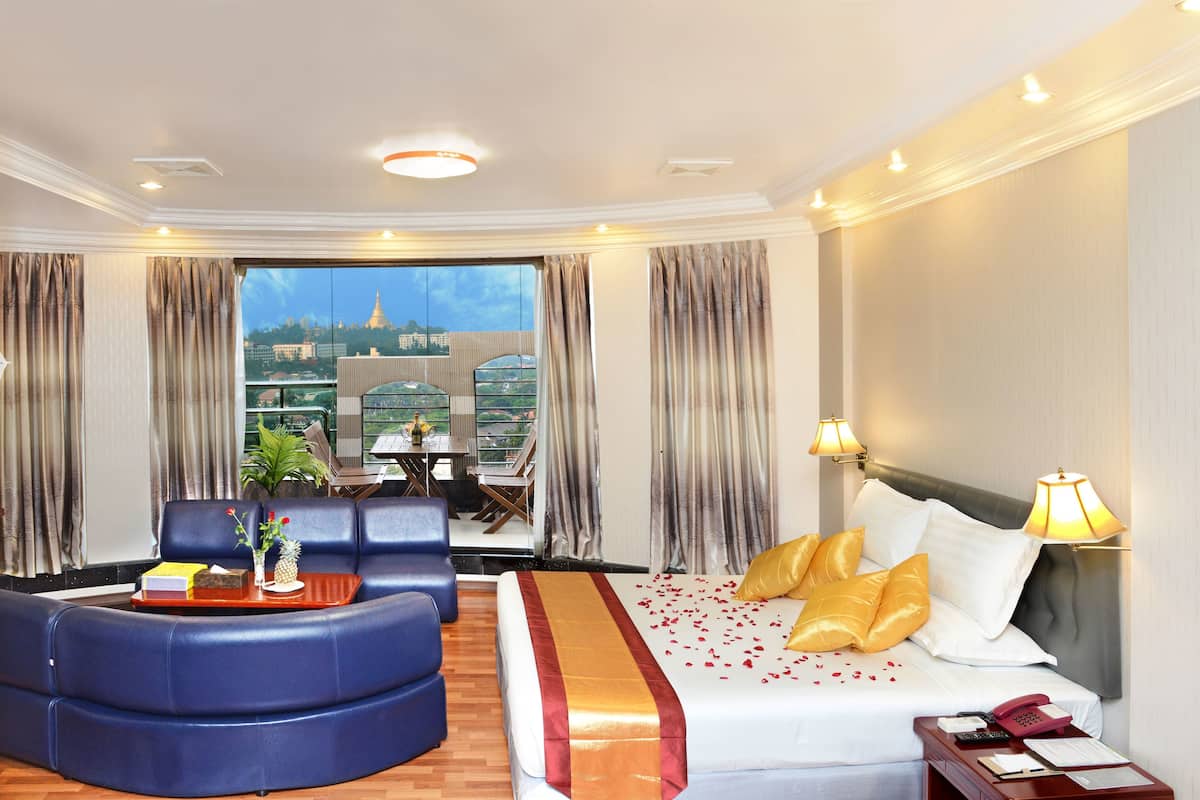 A hotel room with a bed, a curved blue sofa, a dining table with chairs, and a view of a cityscape through large windows.