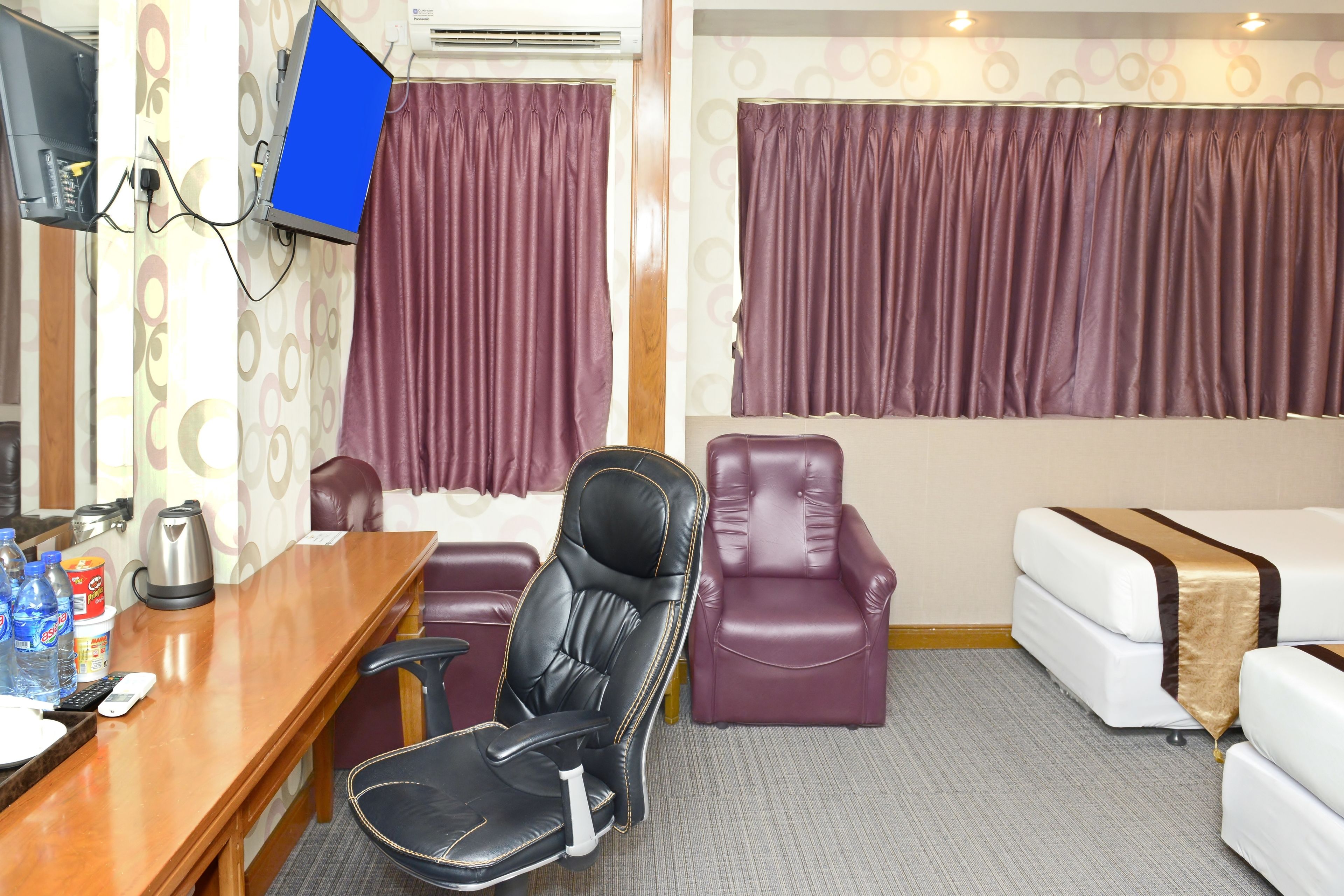 Habitació d'hotel amb escriptori de fusta, una cadira de cuir negre, un sof à de cuir morat i un llit blanc amb detall daurat, juntament amb cortines morades.