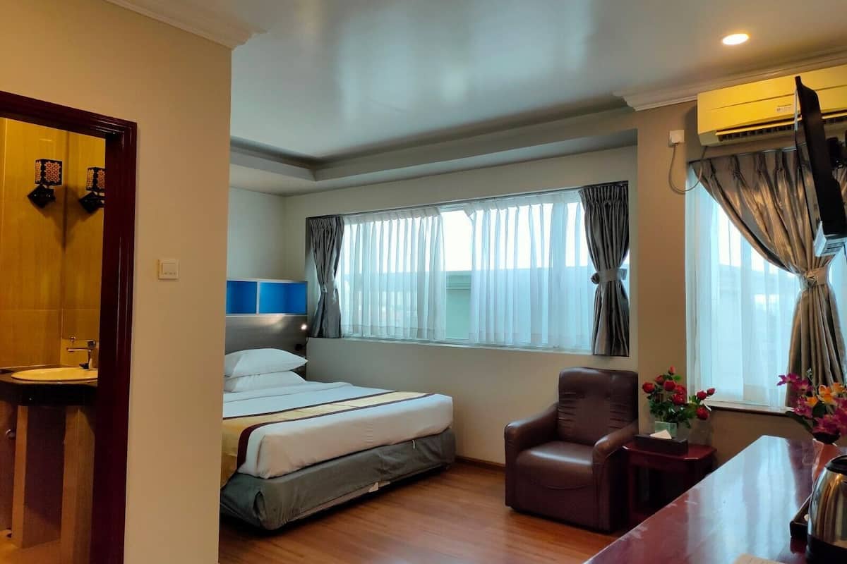 A hotel room with a bed, a chair, a desk, a window with curtains, and an air conditioning unit.