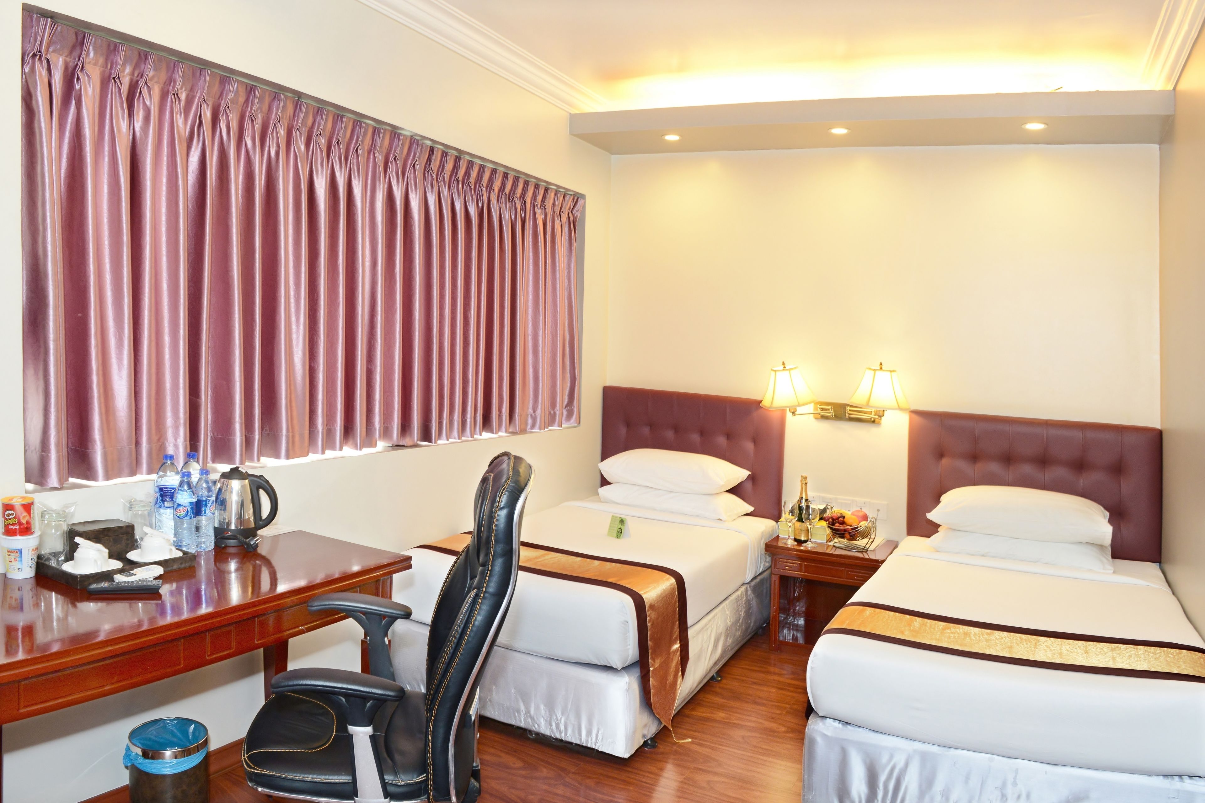 A hotel room with two beds, a desk with a chair, a small table with a lamp, and a window with curtains.