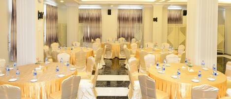 A banquet hall with round tables, chairs, and a central aisle. Tables are set with water bottles and plates.