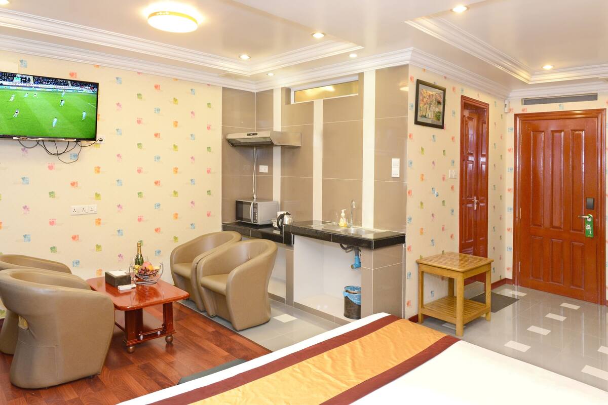 A hotel room with a TV, a sofa, a small table with a bottle and glasses, a chair, a kitchenette, and a wooden door.