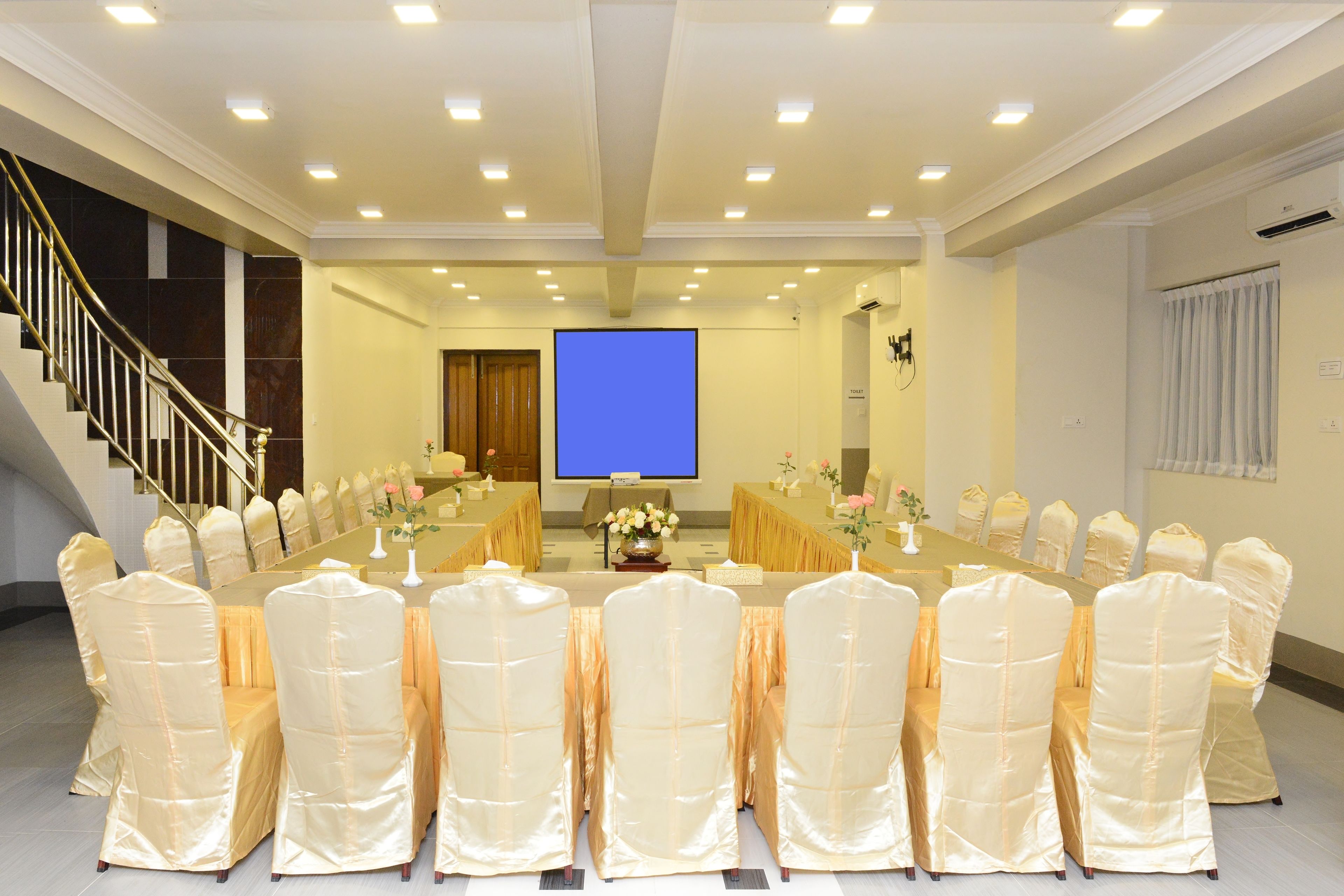 A conference room with a long table and chairs, a projector screen, and a staircase.
