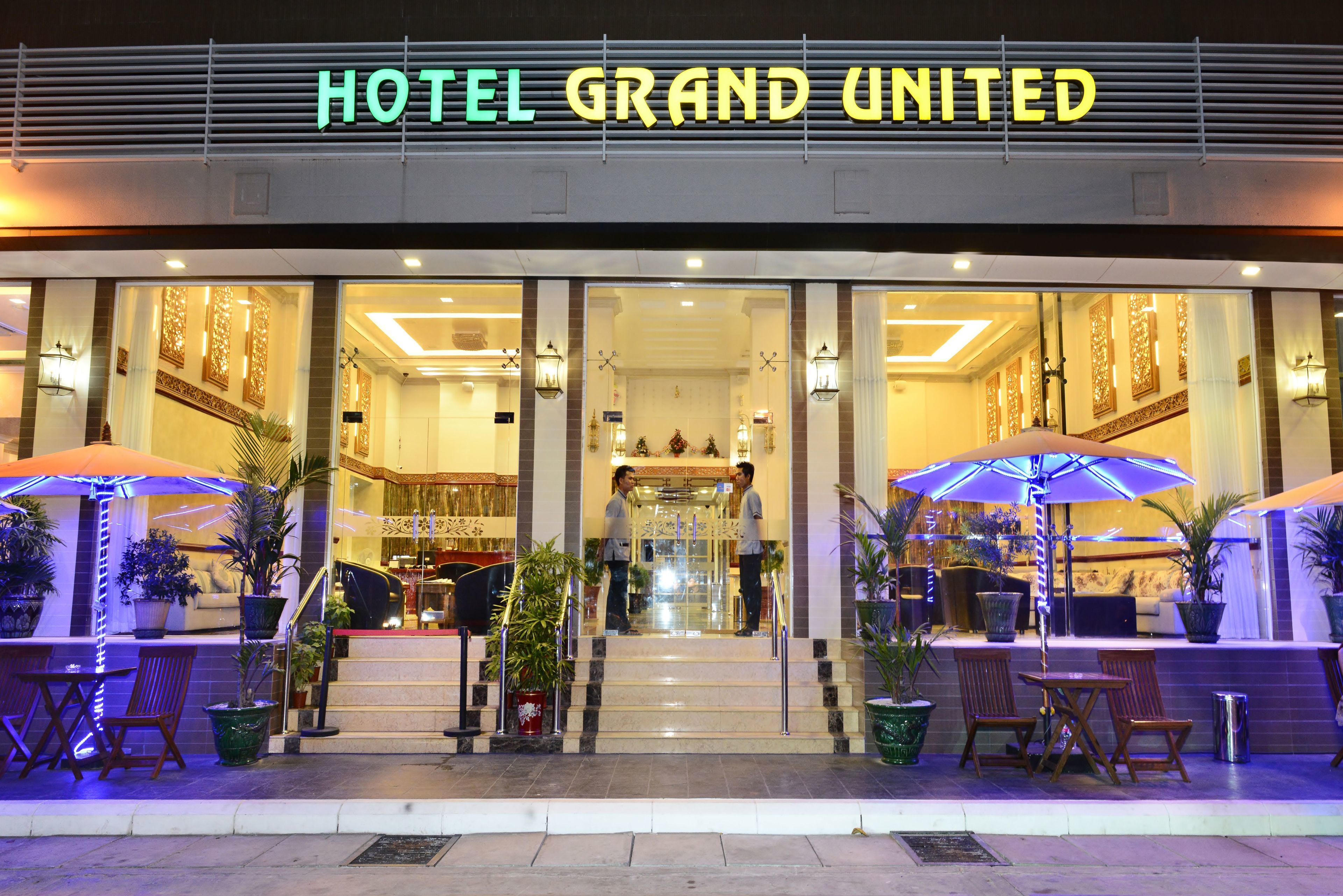The entrance of HOTEL GRAND UNITED with a glass facade, outdoor seating area, and a staircase leading to the lobby.