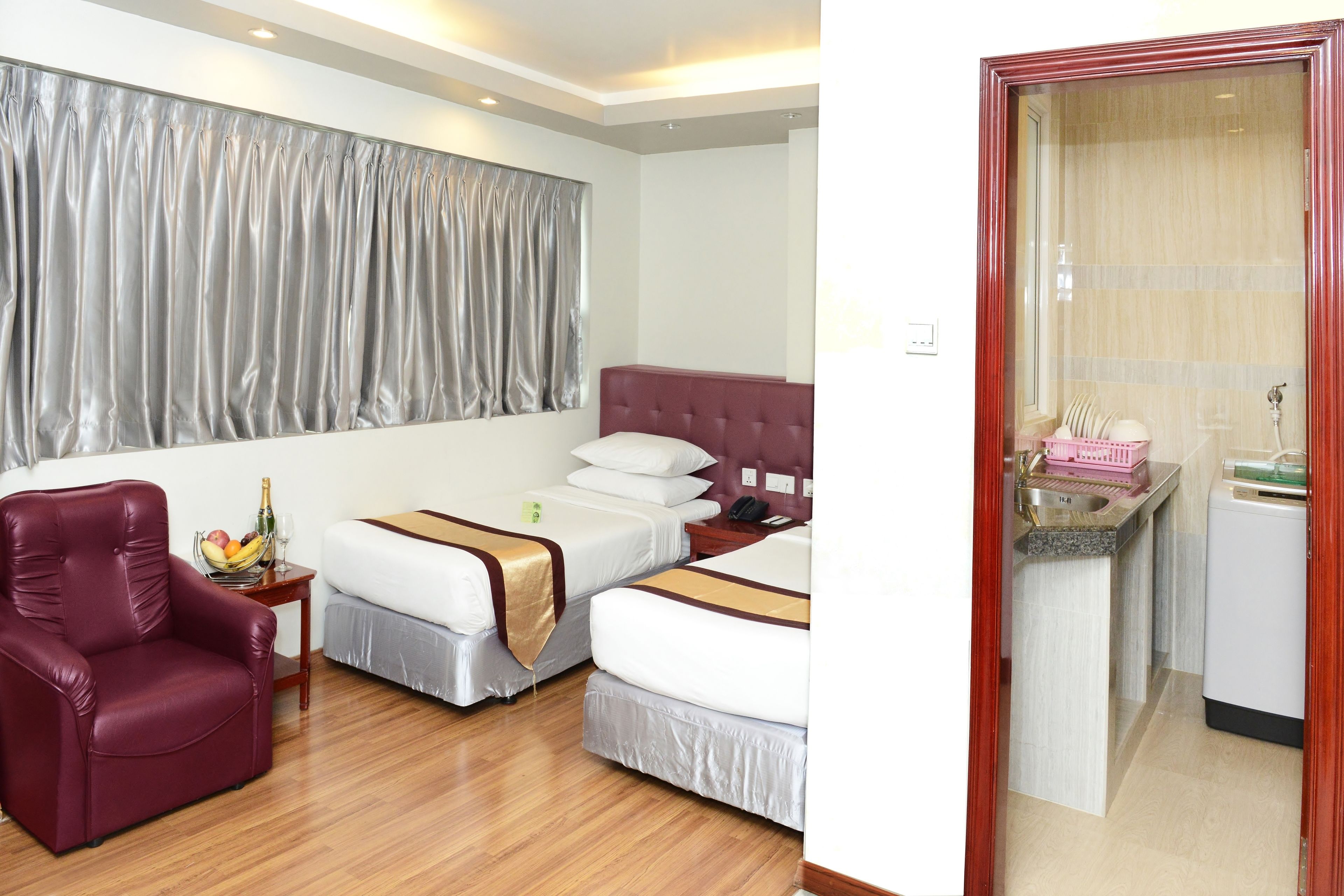Habitación de hotel con dos camas, un sillón rojo, una mesita con frutero y una botella, y vistas a un baño con lavamanos y espejo a través de una puerta.