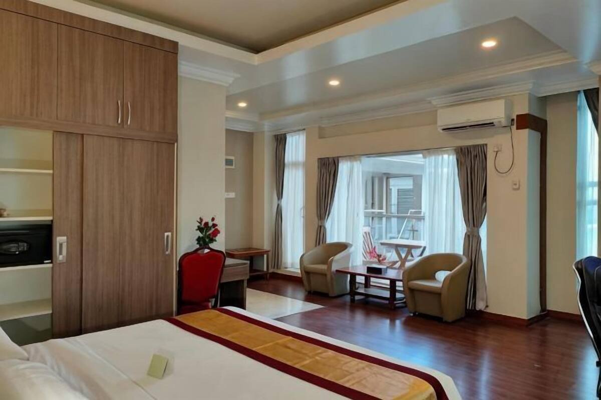 A hotel room with a bed, a desk, two chairs, a wardrobe, and an air conditioning unit.