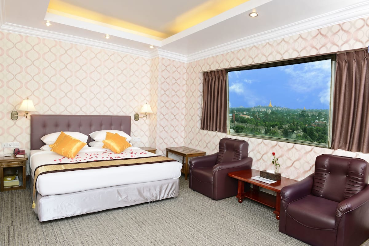 A hotel room with a large bed, two armchairs, a small table, and a view of a cityscape through a large window.