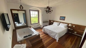 A bedroom with a large bed, a built-in bathtub, a wooden floor, a mirror, a window, and a chandelier. - Hotel Solar das Lajes (Ouro Preto)