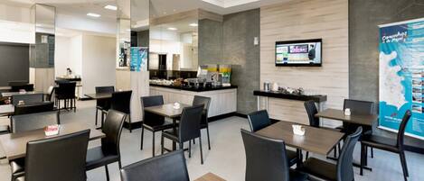 Un comedor de hotel moderno con varias mesas y sillas, una cocina al fondo y un cartel grande en la pared.