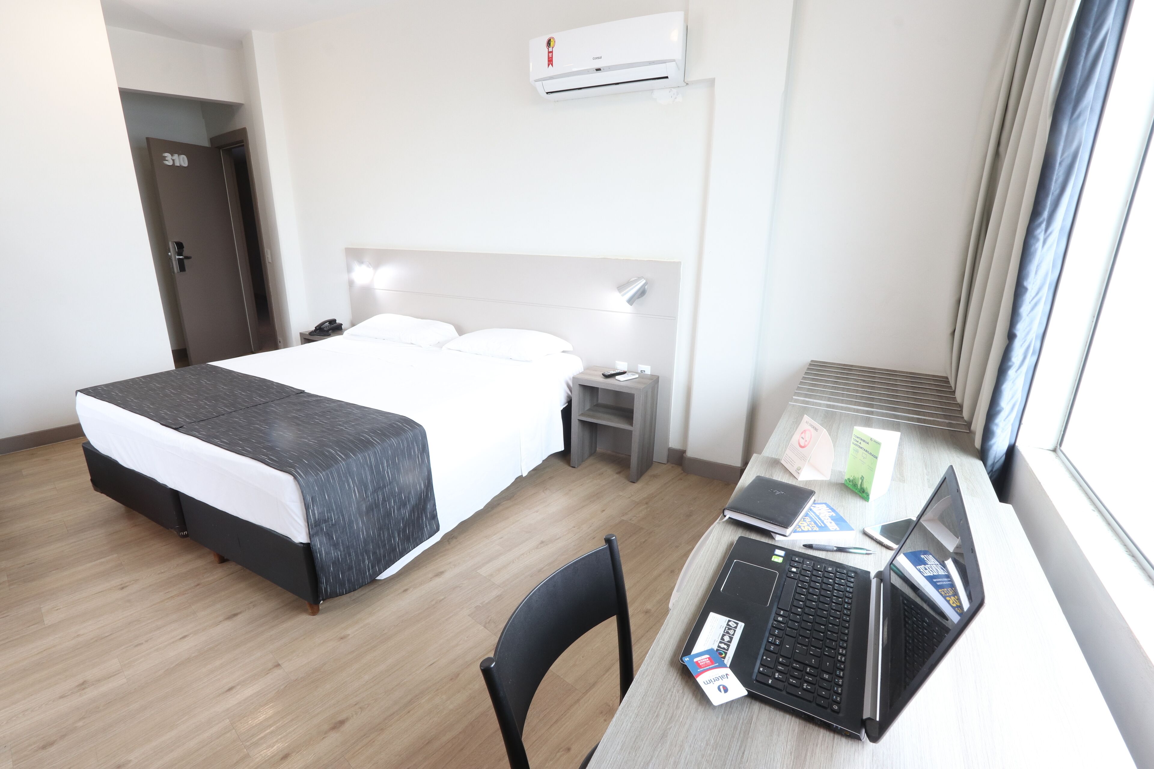 A hotel room with a bed, desk, laptop, and a window.