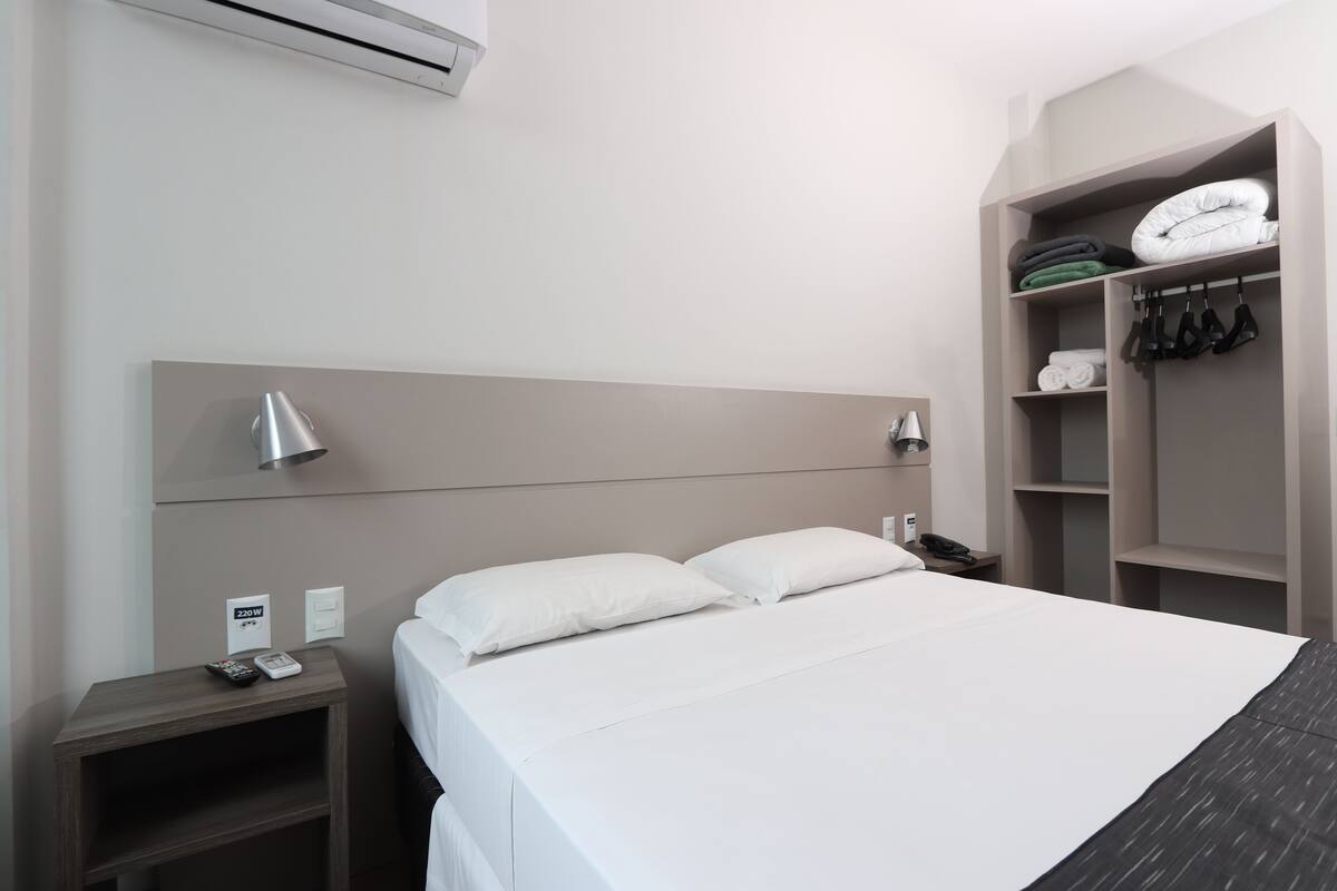 A modern hotel room with a large bed, a bedside table with a lamp, a wardrobe, and a small shelf with folded towels.