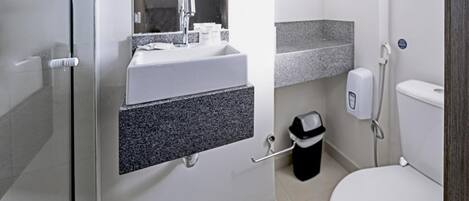 Un baño moderno con cabina de ducha de vidrio, un lavamanos blanco con encimera de granito, un inodoro y un cubo de basura.