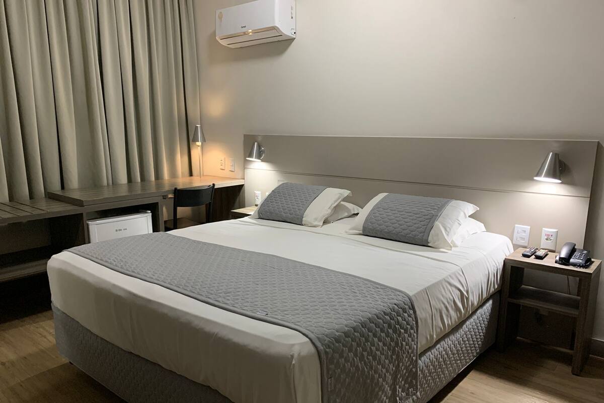 A hotel room with a large bed, bedside tables, a desk, and an air conditioning unit.