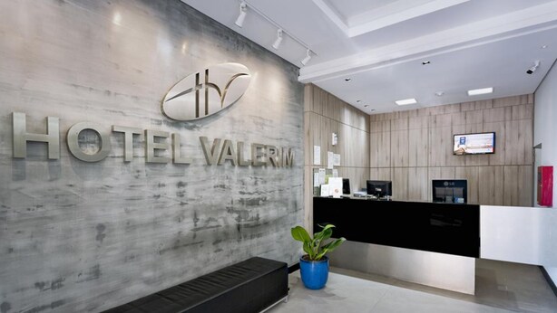 A modern hotel reception area with a marble wall, a reception desk, a bench, and a potted plant. - Hotel Valerim Itajaí (Itajai)