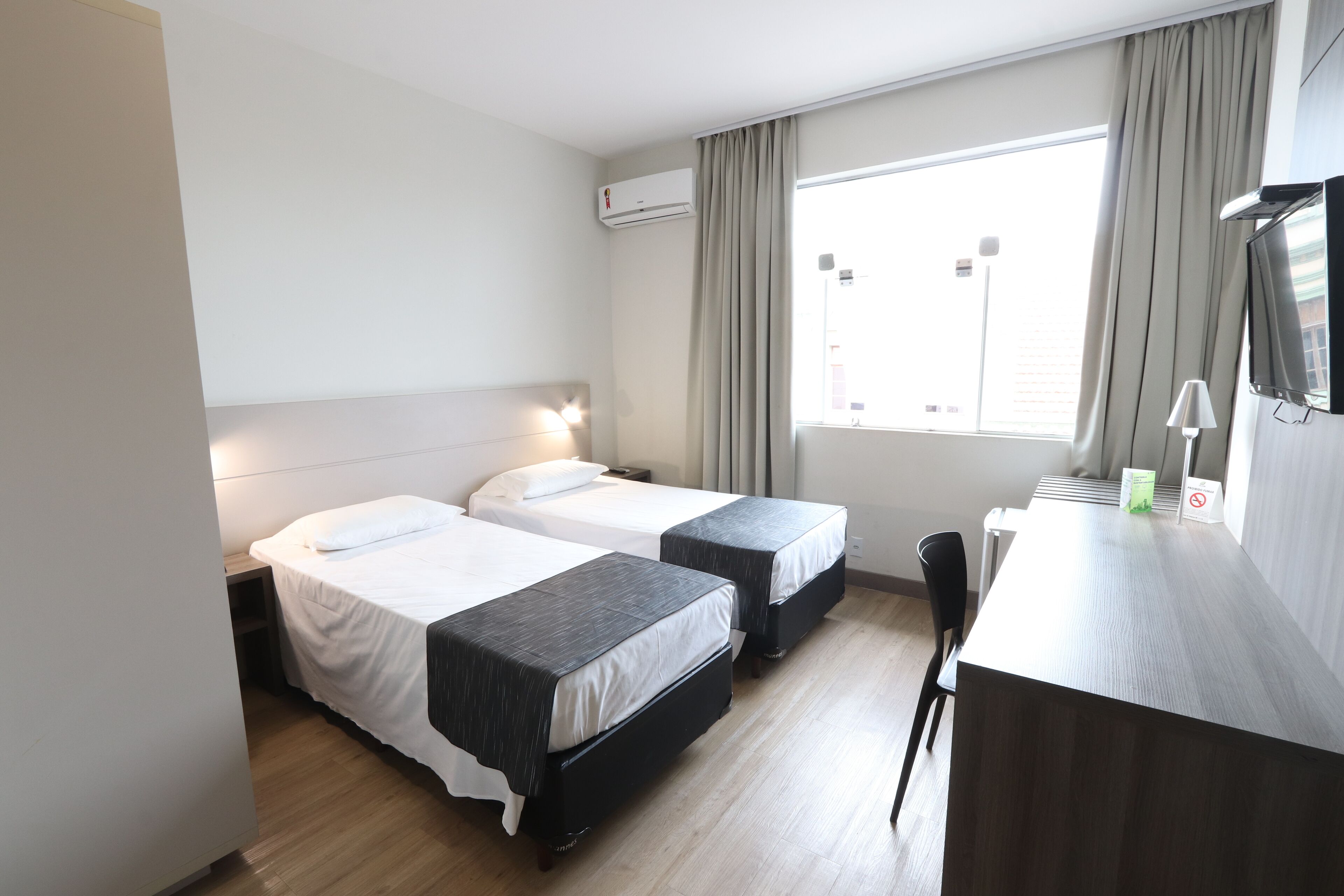 Ett hotellrum med två separata sängar, ett skrivbord med en stol, ett fönster med gardiner och en luftkonditioneringsanläggning.