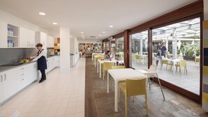 A modern kitchen with white cabinets, a person preparing food, and a dining area with yellow chairs and tables. - Ethra Reserve - Valentino 4* (Castellaneta Marina)