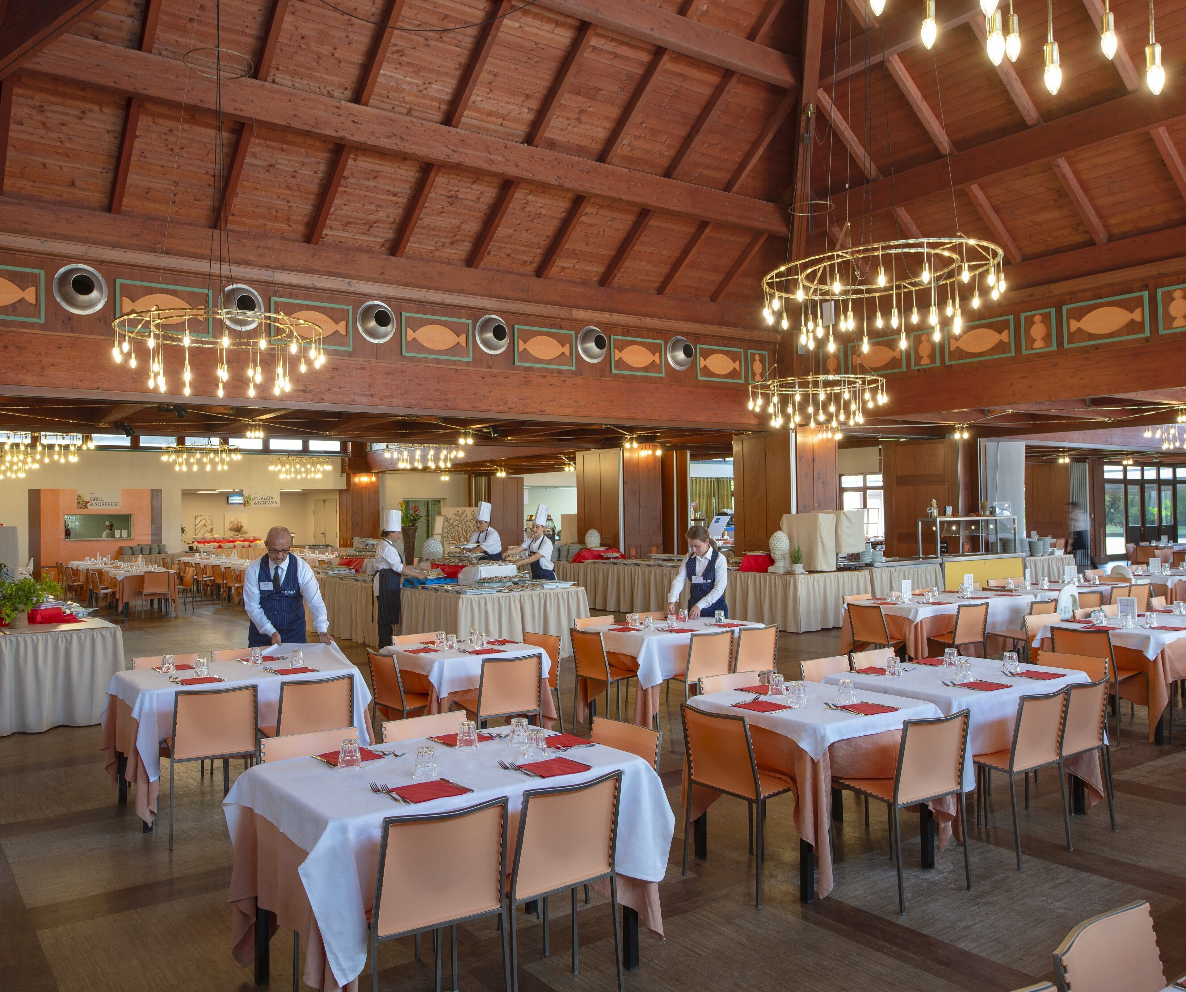 Een ruime eetzaal met meerdere gedekte tafels voor gasten, een hoog houten plafond en moderne kroonluchters.