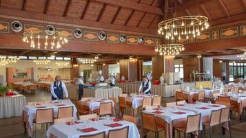 Een ruime eetzaal met meerdere gedekte tafels voor gasten, een hoog houten plafond en moderne kroonluchters.
