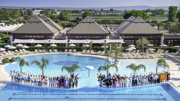 A large pool with lounge chairs and umbrellas, surrounded by a landscaped area with trees and a view of distant buildings. - Ethra Reserve - Valentino 4* (Castellaneta Marina)