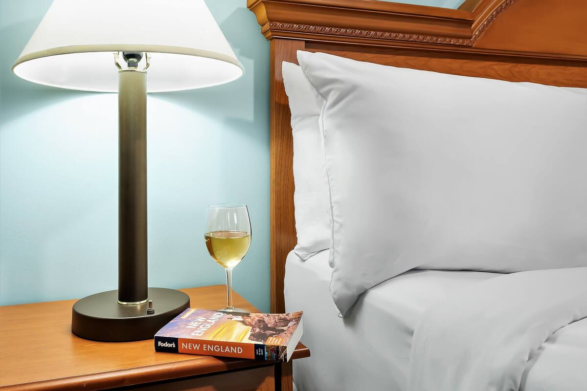 Una cama bien hecha con sábanas blancas, cabecera de madera, una lámpara de noche y una copa de vino blanco sobre una mesita de noche de madera.