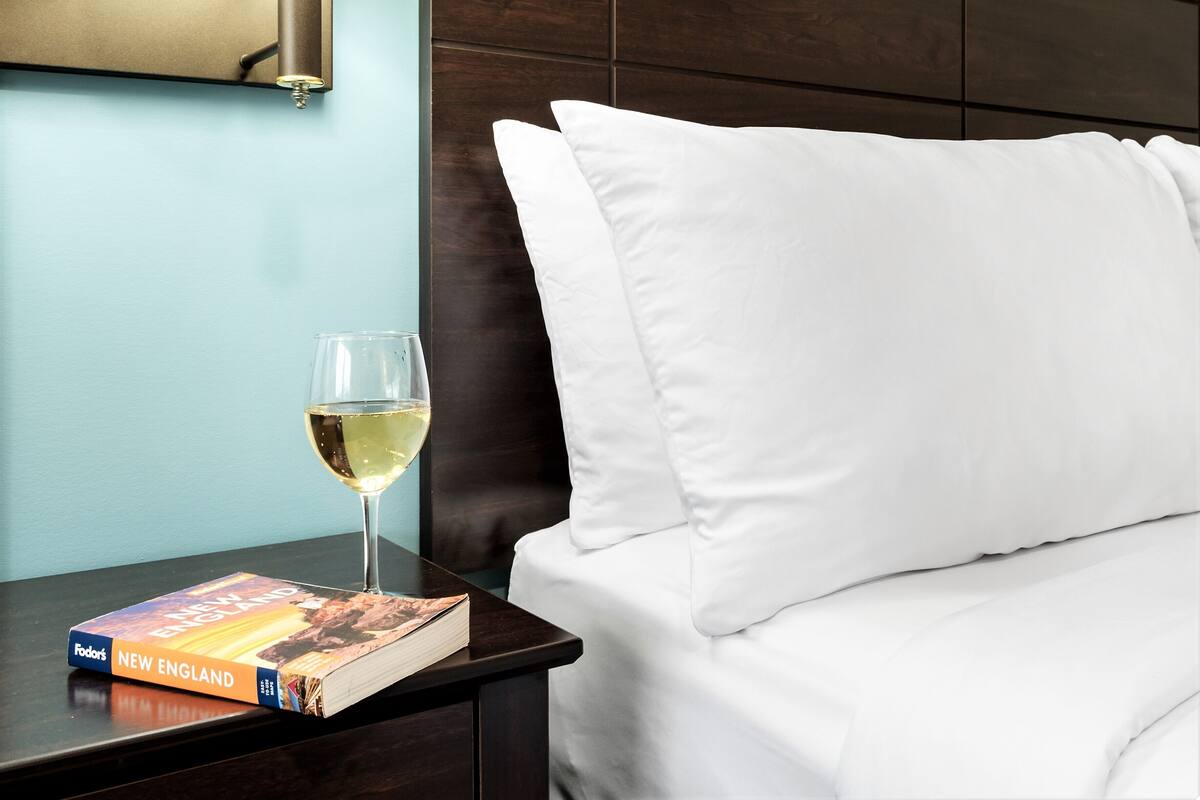 Una cama bien hecha con sábanas blancas, una mesita de noche con un libro titulado 