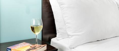 Un lit bien fait, avec du linge de lit blanc, une table de chevet sur laquelle repose un livre intitulé « Nouvelle-Angleterre » et un verre de vin blanc, ainsi qu’une lampe fixée au mur.