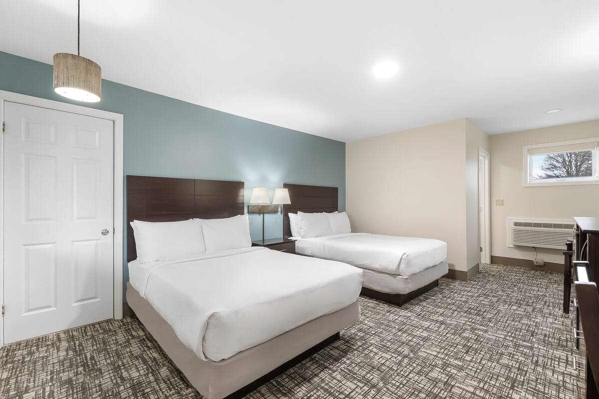 Habitación de hotel con dos camas, cabecero de madera, alfombra estampada, puerta blanca y ventana con persianas.