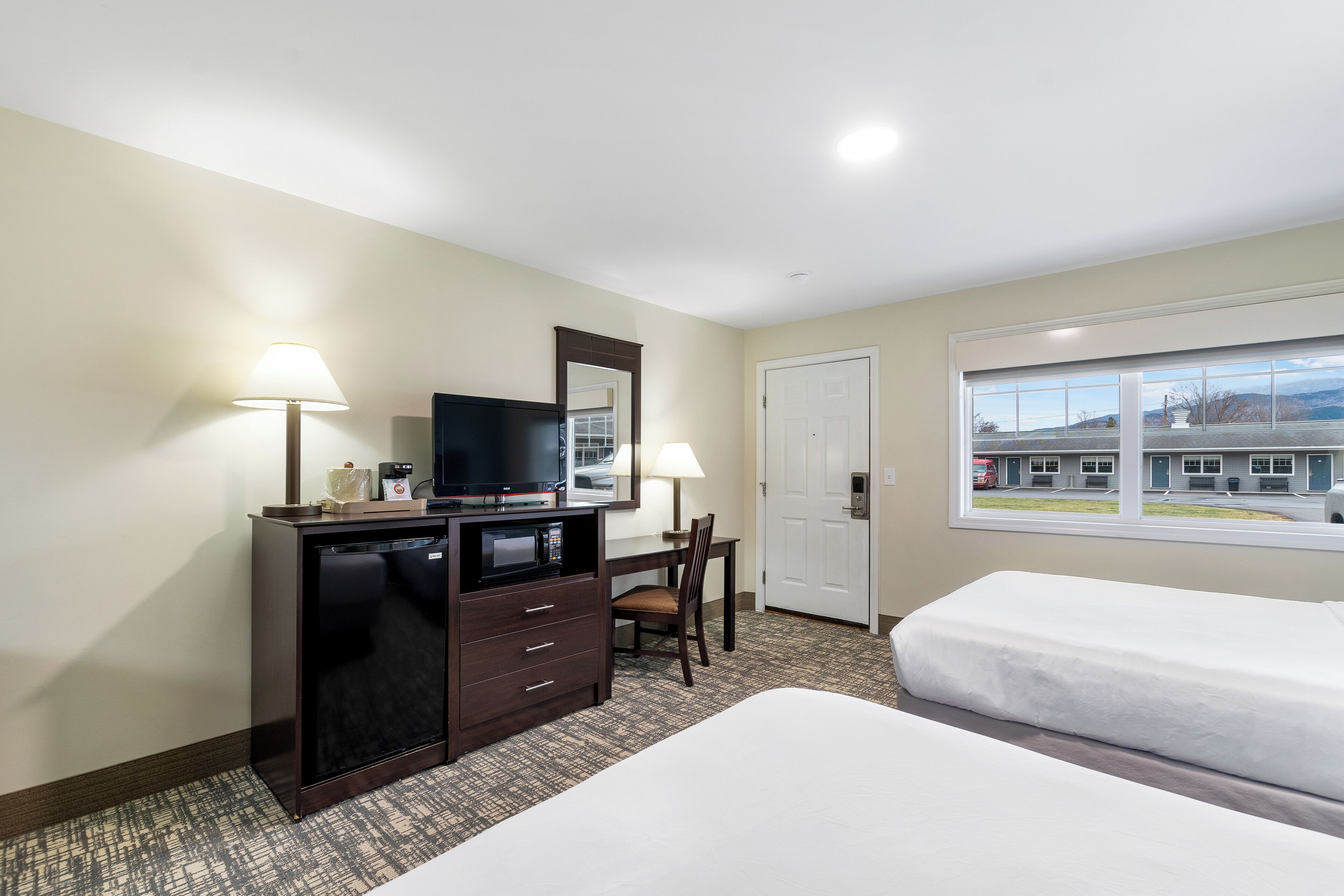 A hotel room with two beds, a desk with a television, a dresser, and a window with a view of a building and parking lot.