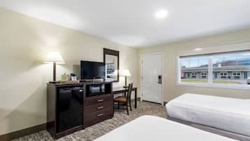 A hotel room with two beds, a desk with a television, a dresser, and a window with a view of a building and parking lot.